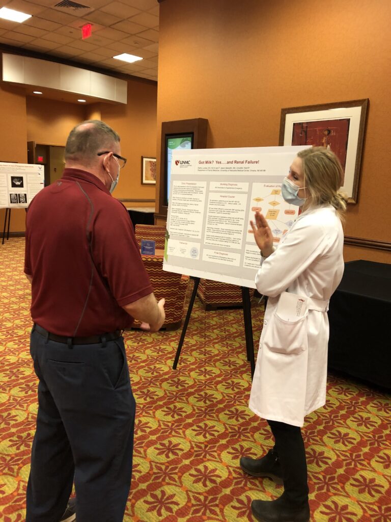 A woman in a white coat explains a scientific poster to a man in a red shirt. Both wear masks, standing on patterned carpet in a warmly lit room.