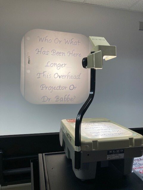 An overhead projector displays the humorous text, "Who or what has been here longer, this overhead projector or Dr. Babbe?" on a wall.