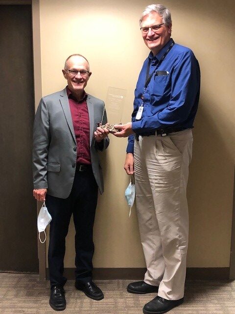 Two men smile while holding an award. One wears a gray blazer, the other a blue shirt. They appear happy. Masks hang from their hands.