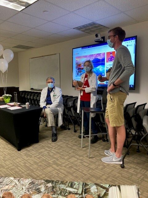 Three people in a room, all wearing masks. Two, in lab coats, engage with a standing man in casual attire. A presentation is projected behind them.