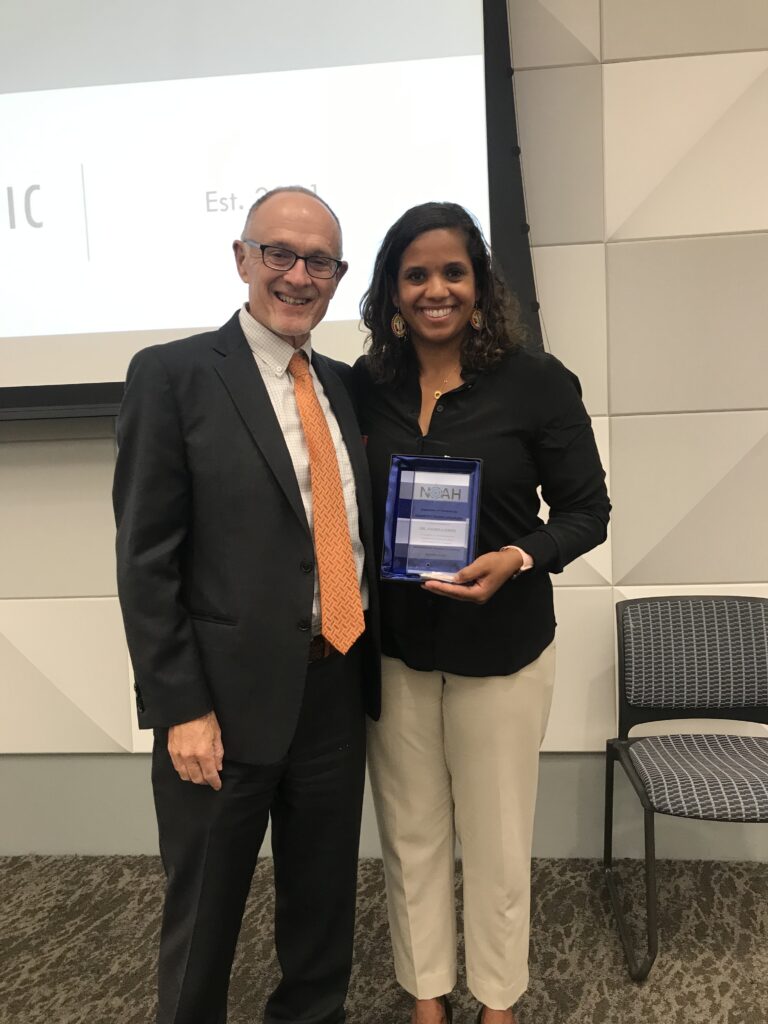 Two smiling individuals stand in a modern room. The woman holds an award plaque.