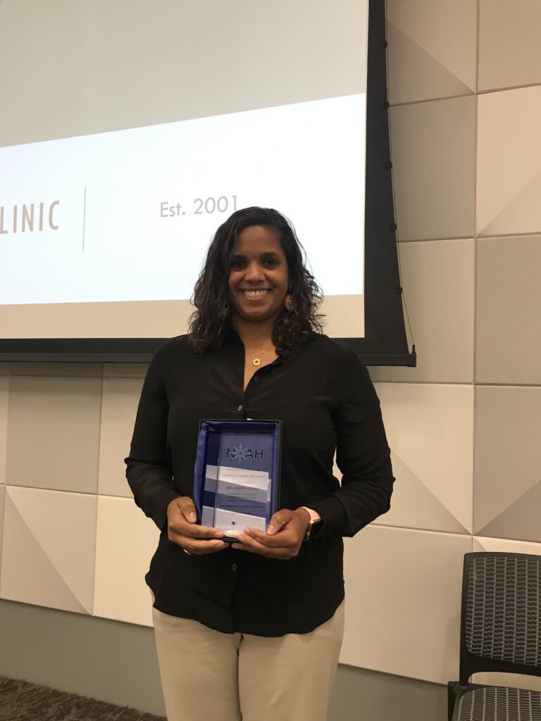 A person in a black shirt smiles while holding an award plaque. They stand in a room with a presentation
