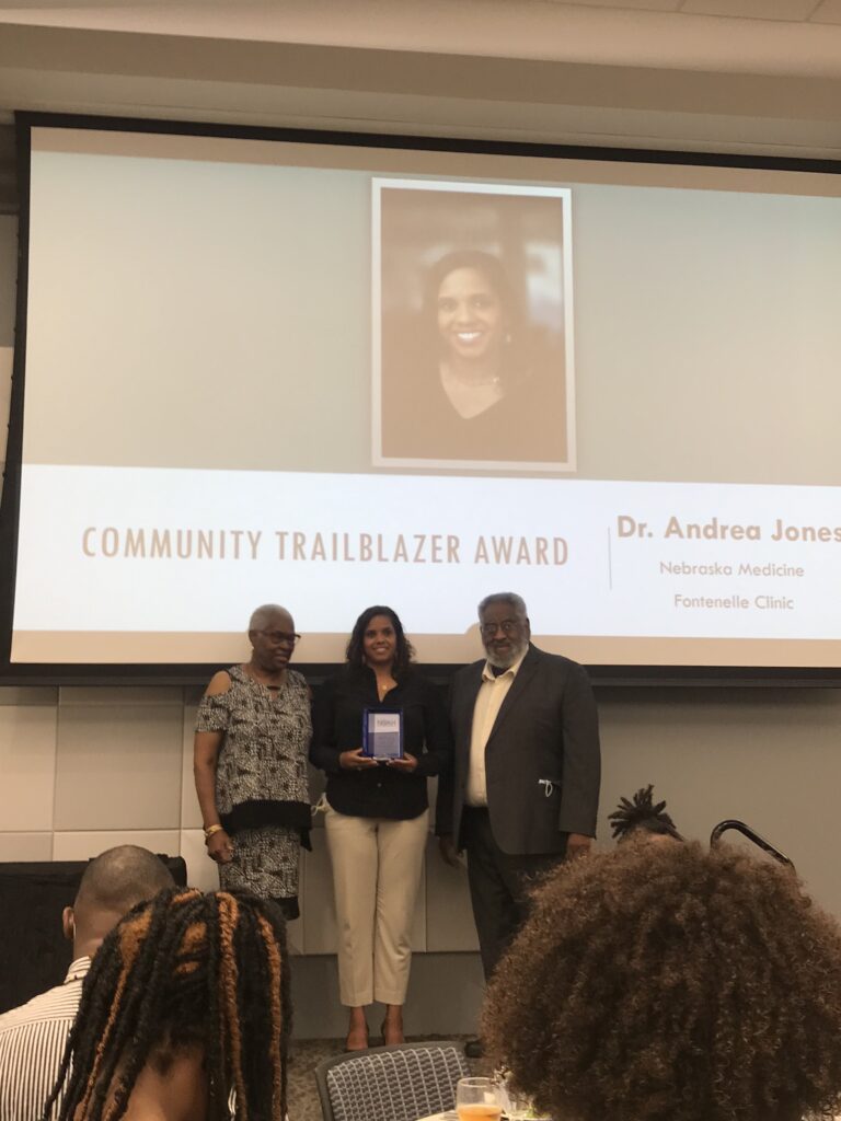 Three people stand in front of a projected screen displaying Community Trailblazer Award and a photo. The middle person holds a blue award, smiling.