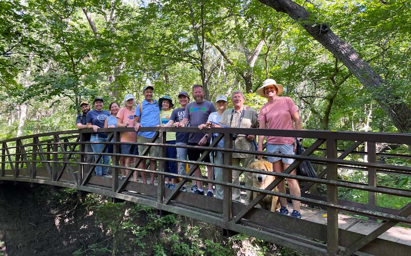 Group of UNMC faculty, spouses, and a dog standing on a bridge in a forest