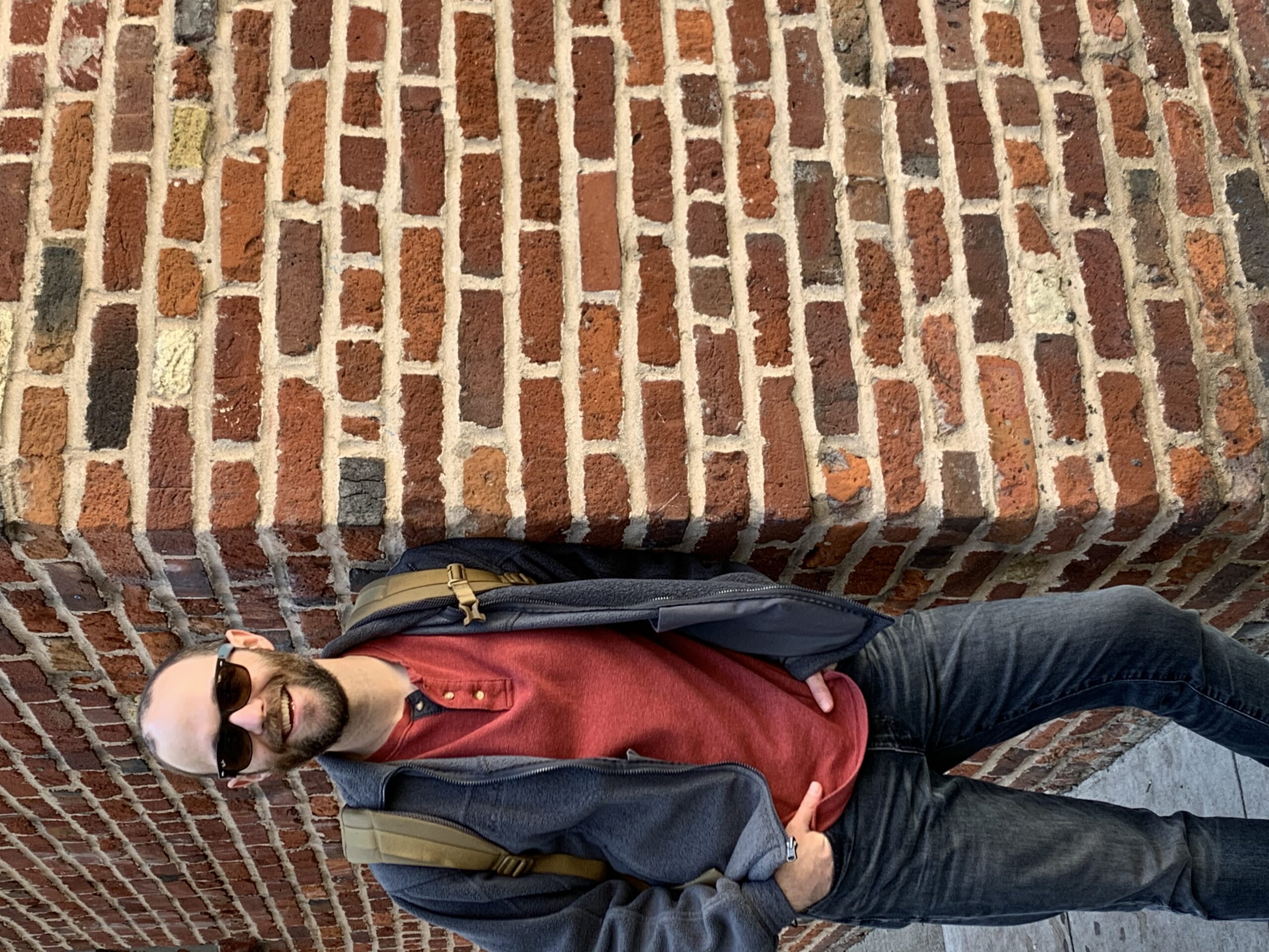 Image of ID fellow Dr. Bryan Walker wearing a red shirt and blue jacket and dark blue jeans and sunglasses leaning against a brick wall