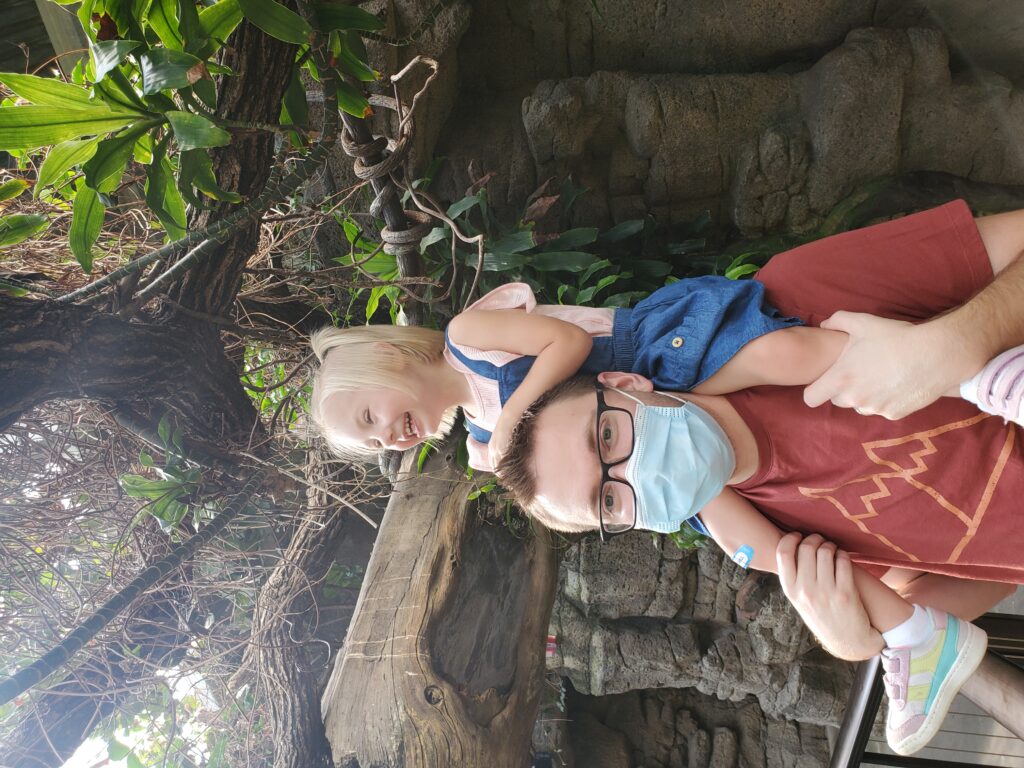 Jeremy wearing a mask with his daughter on his shoulders laughing underneath a tree