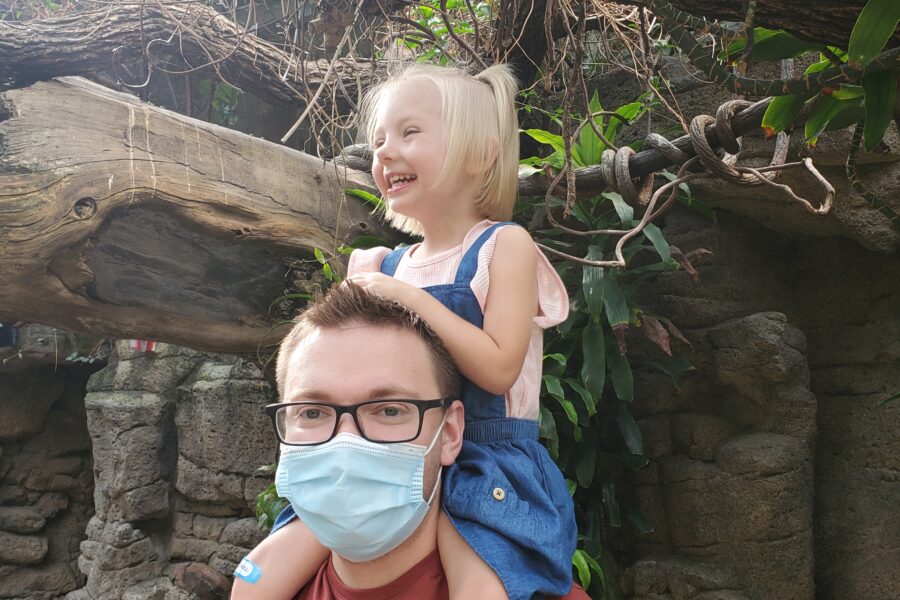 Jeremy wearing a mask with his daughter on his shoulders laughing underneath a tree