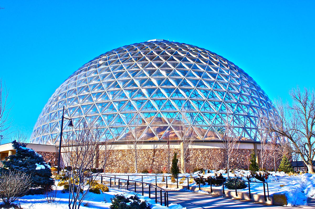 A large geodesic dome structure with a glass and metal framework, set against a bright blue sky. The dome is surrounded by a landscaped area with trees, shrubs, and a walkway. Patches of snow cover the ground, indicating a winter setting. The structure appears to be part of a public space, possibly a botanical garden or zoo exhibit