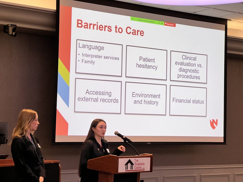 Two women in professional attire are giving a presentation at The Thompson Center. One of them is speaking at a podium while the other stands beside her. A large projection screen behind them displays a slide titled 'Barriers to Care,' listing factors such as language, patient hesitancy, clinical evaluation vs. diagnostic procedures, accessing external records, environment and history, and financial status. The slide features a red and white design with a university logo in the bottom right corner. The room has dim lighting, and the presentation is being shared via Zoom, as indicated by a green screen-sharing bar at the top