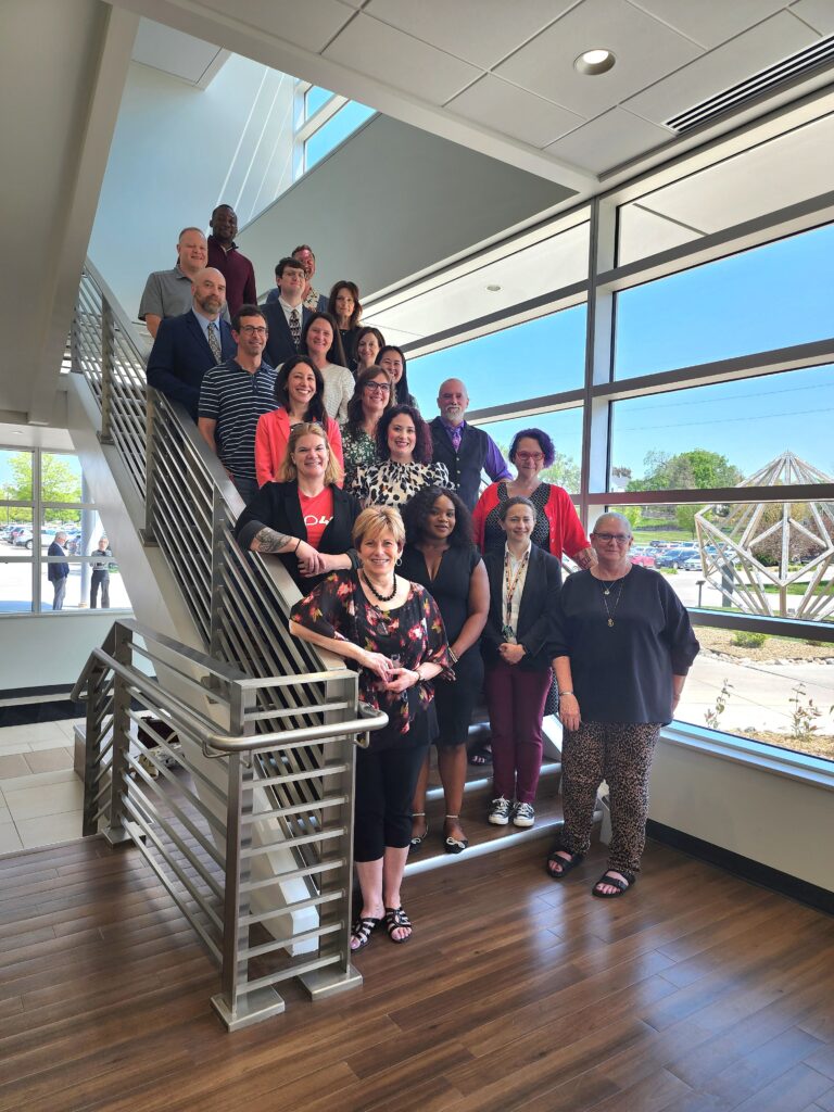 20 people standing on stairs dressed professionally, smiling. in the background outside is a bright day with a blue sky.