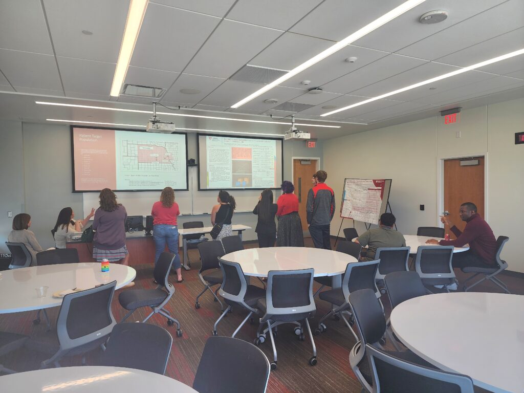 Image depicting several people in a classroom setting standing in front of two presentation screens with white backgrounds, text not legible. some people are seated in chairs around a table.
