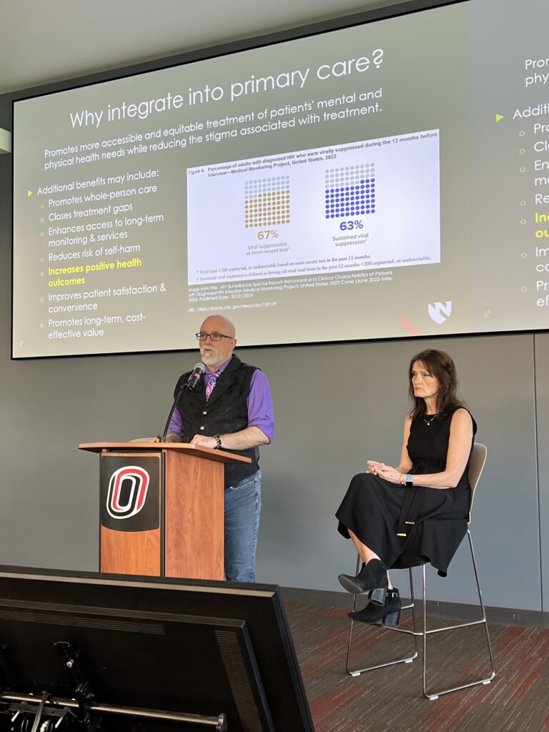 presentation screen with dark background and white text, in the center is a blurry image with white background and red and blue dots depicting program participation. In the foreground are two presenters dressed professionally, one standing at a brown podium and the other sitting in a chair to his left.