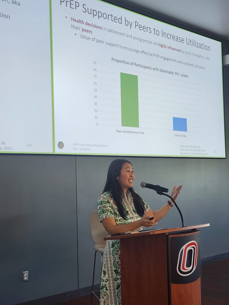 In the foreground, presenter dressed in green and white dress standing in front a brown podium with microphone. in background a presentation screen with white background, black lettering at the top saying "PrEP supported by Peers to Increase Utilization". Graph with blue and green bars in center of screen. remainder of text not legible in photo.