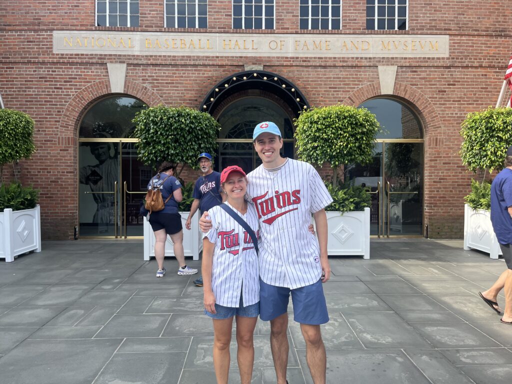 two people standing wearing twins jerseys in foreground. background building with other people