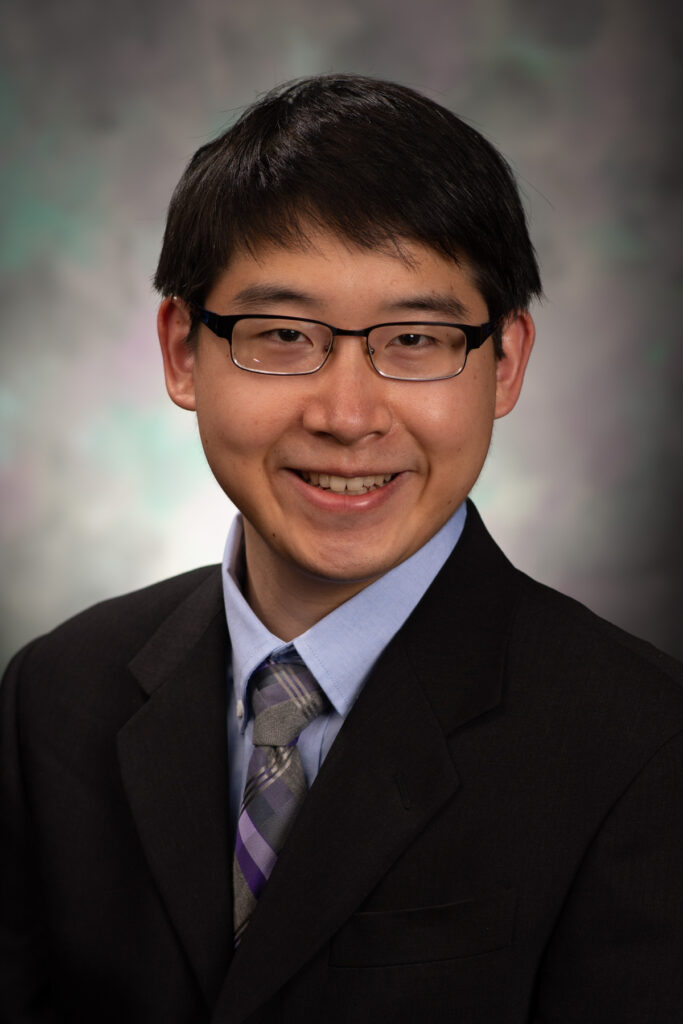 portrait photo. subject wearing black suit with blue dress shirt and grey tie. wearing glasses and smiling.