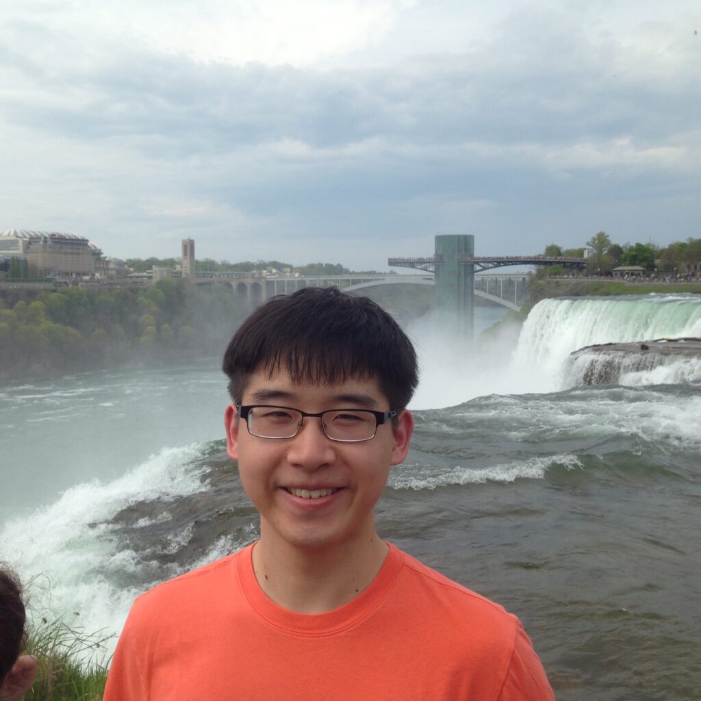 background: waterfalls
foreground: subject standing wearing orange t shirt, wearing glasses, smiling
