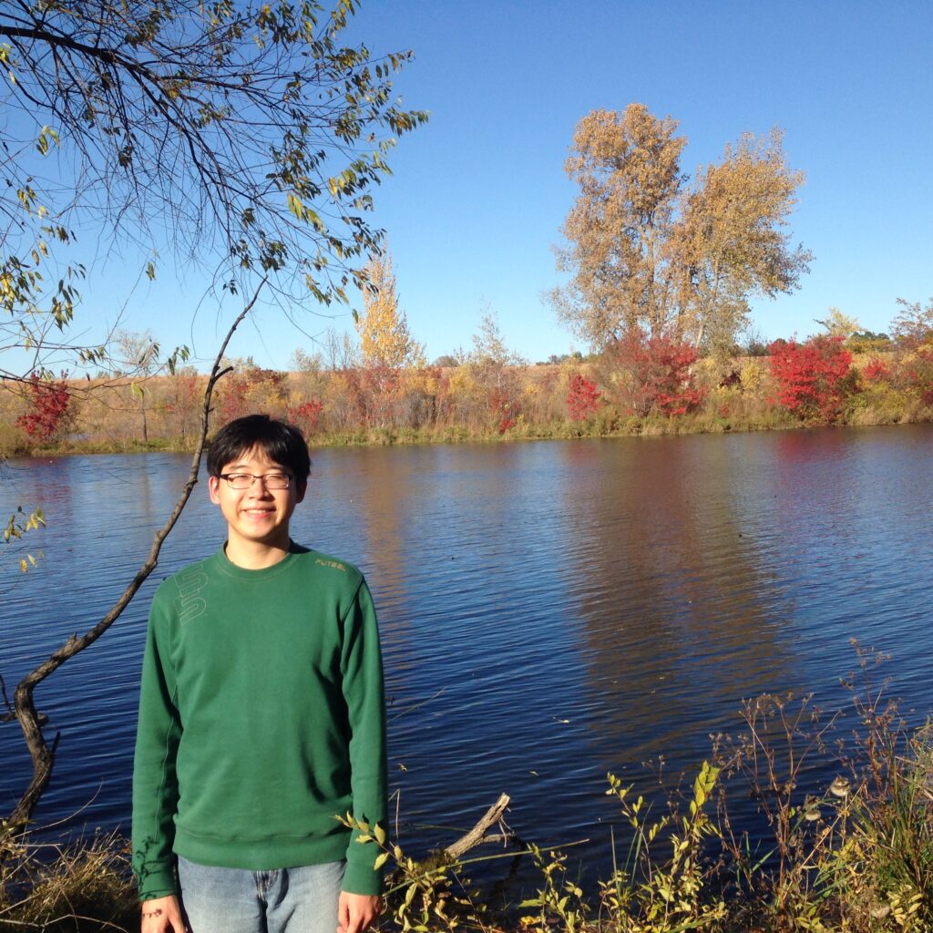 background: lake with fall foliage
foreground: subject standing wearing green long-sleeved t shirt and grey jeans, wearing glasses, smiling