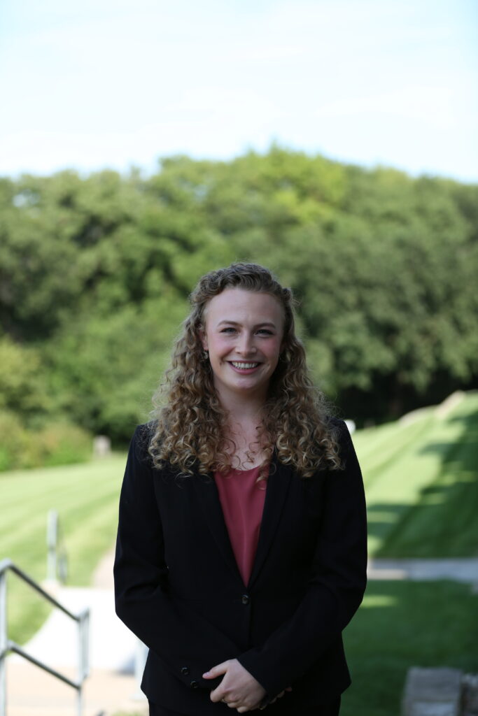 headshot of a smiling woman wearing a black blazer and red shirt. in the background are green trees and lawn.