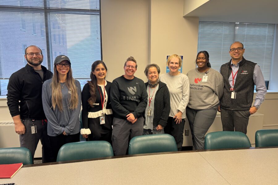 Seven people standing in a conference room behind a long table with teal chairs&period; They wear casual and business-casual clothing with ID badge lanyards&period; The background shows two large windows with blinds&comma; light-colored walls&comma; and a small colorful poster between the windows&period;