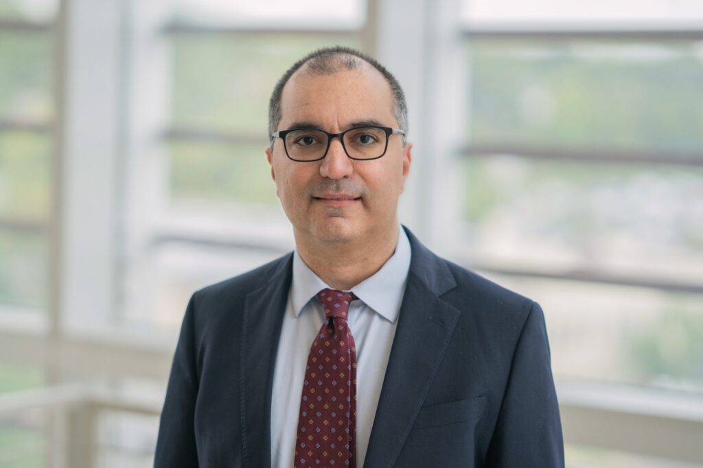 Headshot of a man wearing a dark suit, red tie, and glasses on a lighted background
