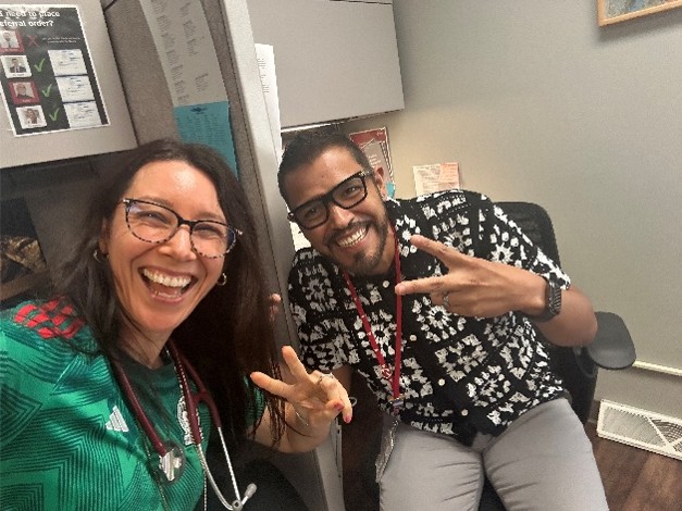 Two people posing for a selfie in an office, both making peace signs and wearing ID badges. One person wears a green Mexico soccer jersey, and office cubicles are visible in the background.