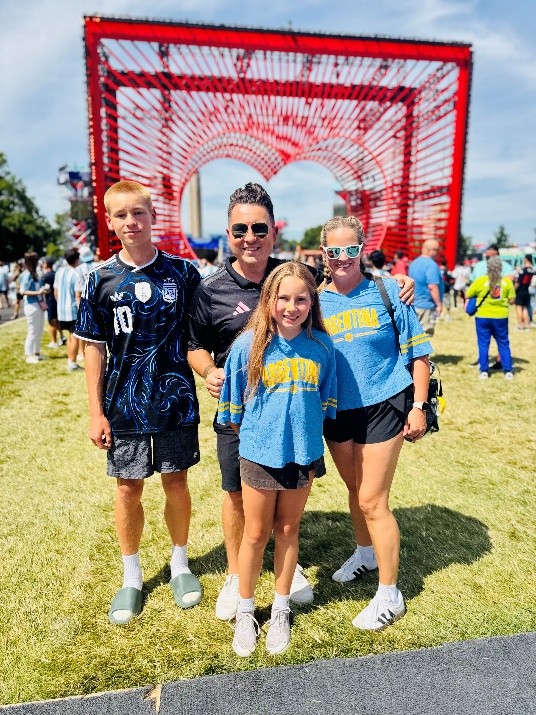 Four people standing together on a grassy field at an outdoor event. Behind them is a large red heart-shaped art installation, with crowds of attendees and a blue sky in the background.