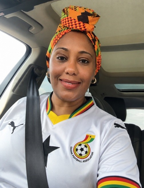Person seated in the front passenger seat of a car wearing a white Ghana national soccer jersey and a patterned orange, yellow, green, and black headwrap. A seatbelt is fastened across the chest, and the vehicle interior is visible in the background.