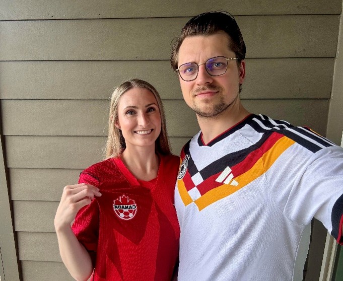 Two people standing side by side in front of a wood-paneled wall, wearing soccer jerseys. One person is wearing a red Canada soccer jersey and holding the team crest toward the camera, while the other is wearing a white Germany national team jersey with black, red, and gold accents.