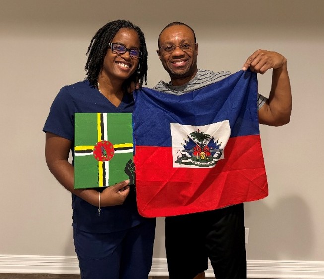 Two people standing indoors against a plain wall, posing with national symbols. One person is holding a painting featuring the flag of Dominica, while the other is holding a Haitian flag.