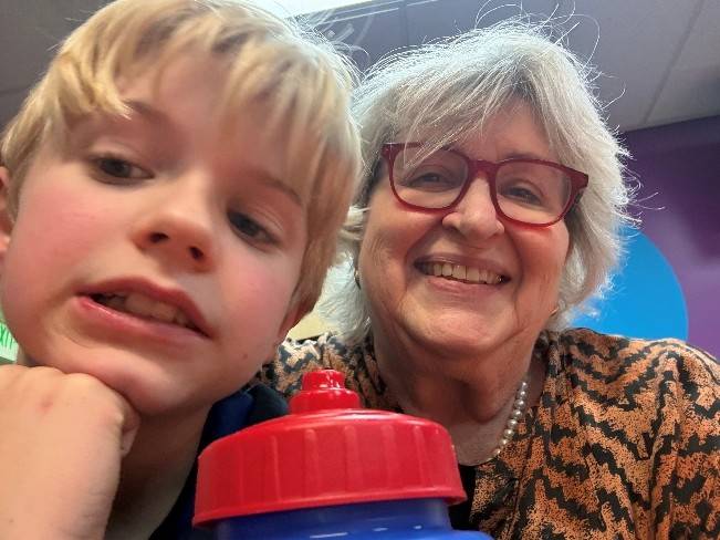 Close-up selfie of a child and an older adult seated indoors at a table. A red and blue water bottle is visible in the foreground, and colorful walls and ceiling lights appear in the background.