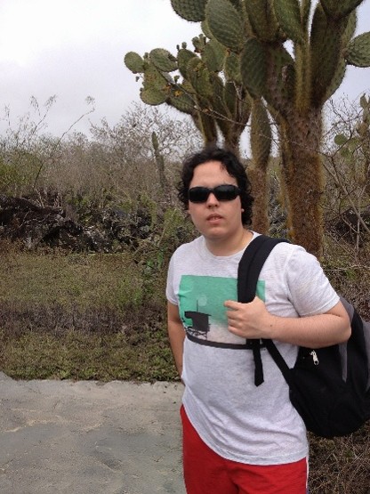 Person wearing sunglasses, a light-colored T-shirt, red shorts, and a backpack standing on a paved path in a dry landscape. Large cactus plants and sparse vegetation are visible behind the person under an overcast sky.