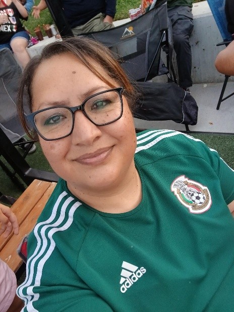 Close-up selfie of a person wearing glasses and a green Mexico national soccer jersey with the team crest and Adidas logo visible. Outdoor seating and other event attendees can be seen in the background.