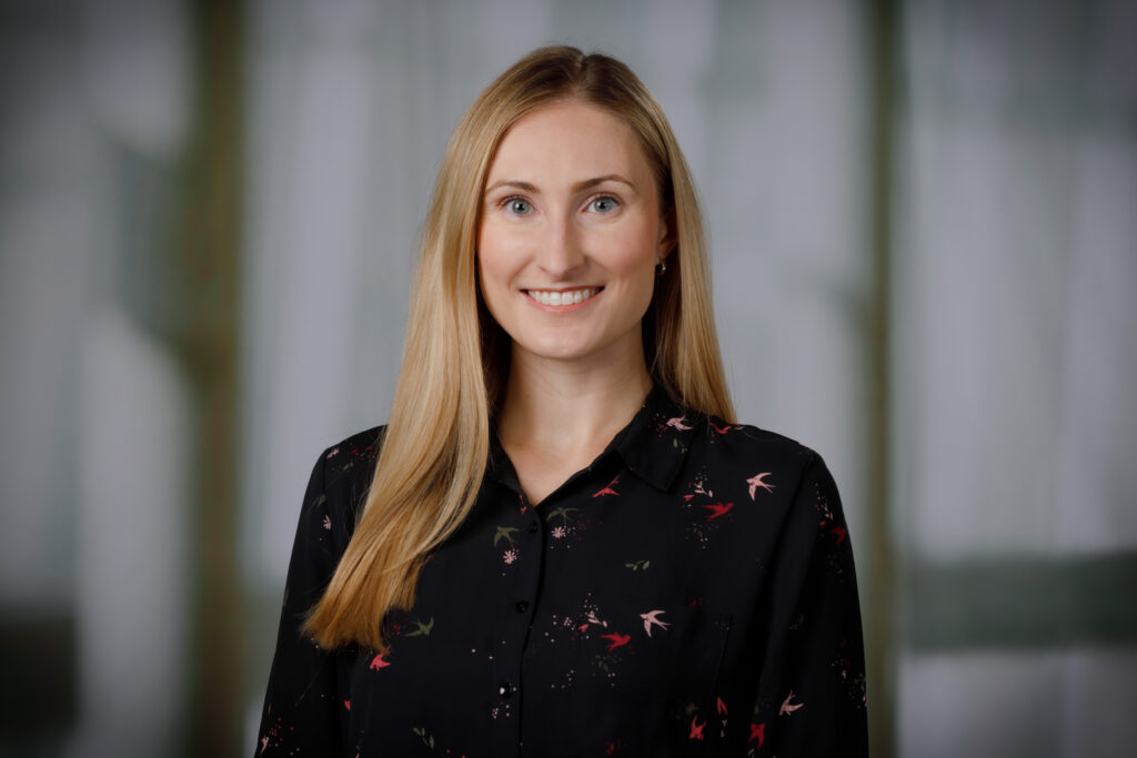 Professional headshot of a person with long hair wearing a dark floral-patterned blouse, photographed against a softly blurred indoor background