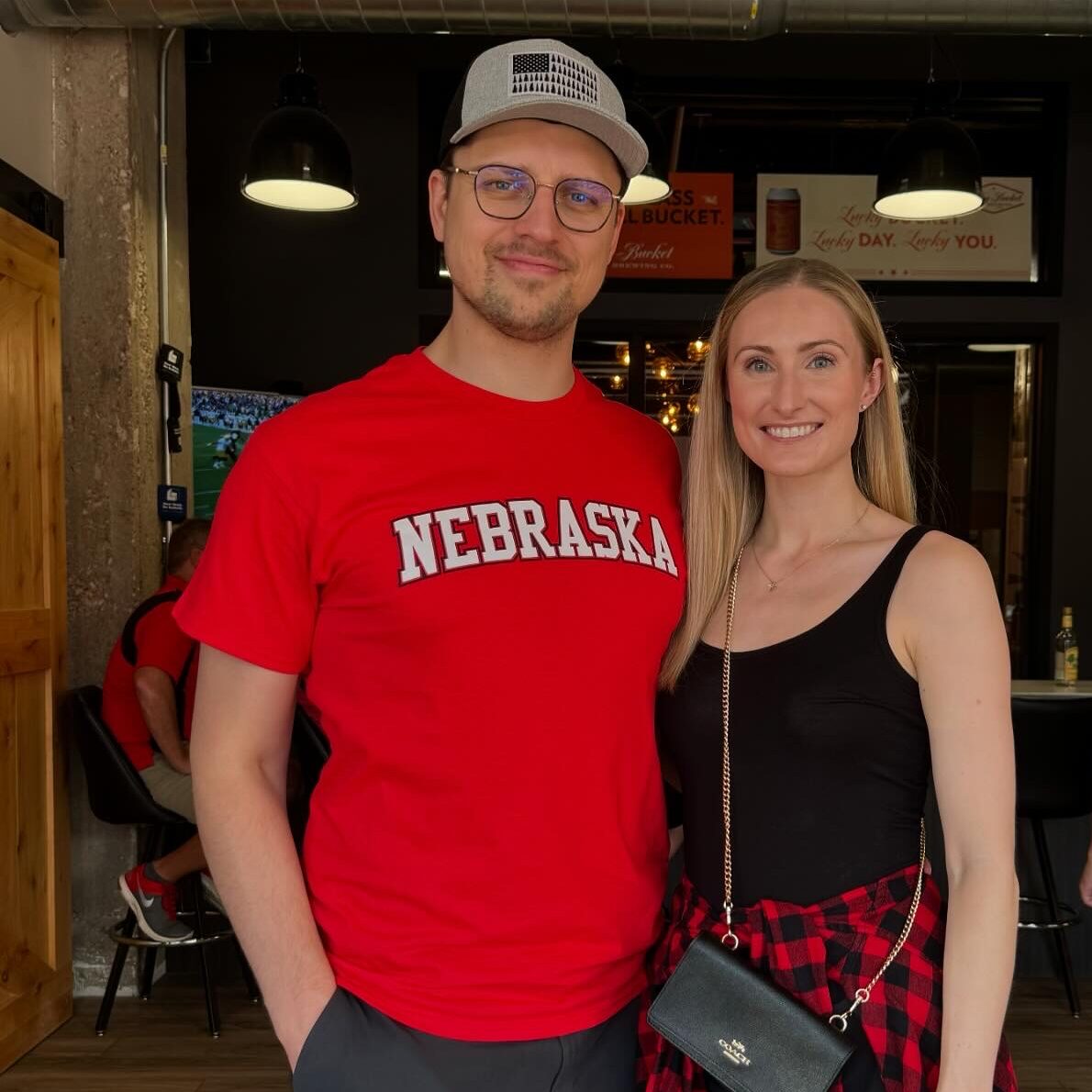 Two people posing indoors at a casual gathering. One person wears a red Nebraska T-shirt, and the other wears a black outfit with a red plaid shirt tied around the waist