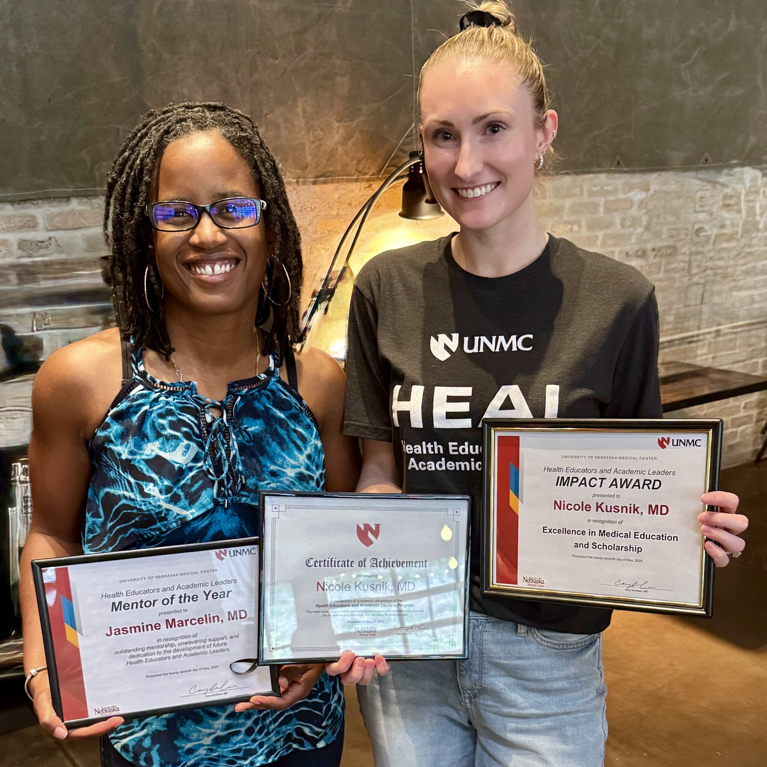 Two award recipients pose with framed UNMC certificates and an impact award during a recognition event celebrating excellence in health professions education.