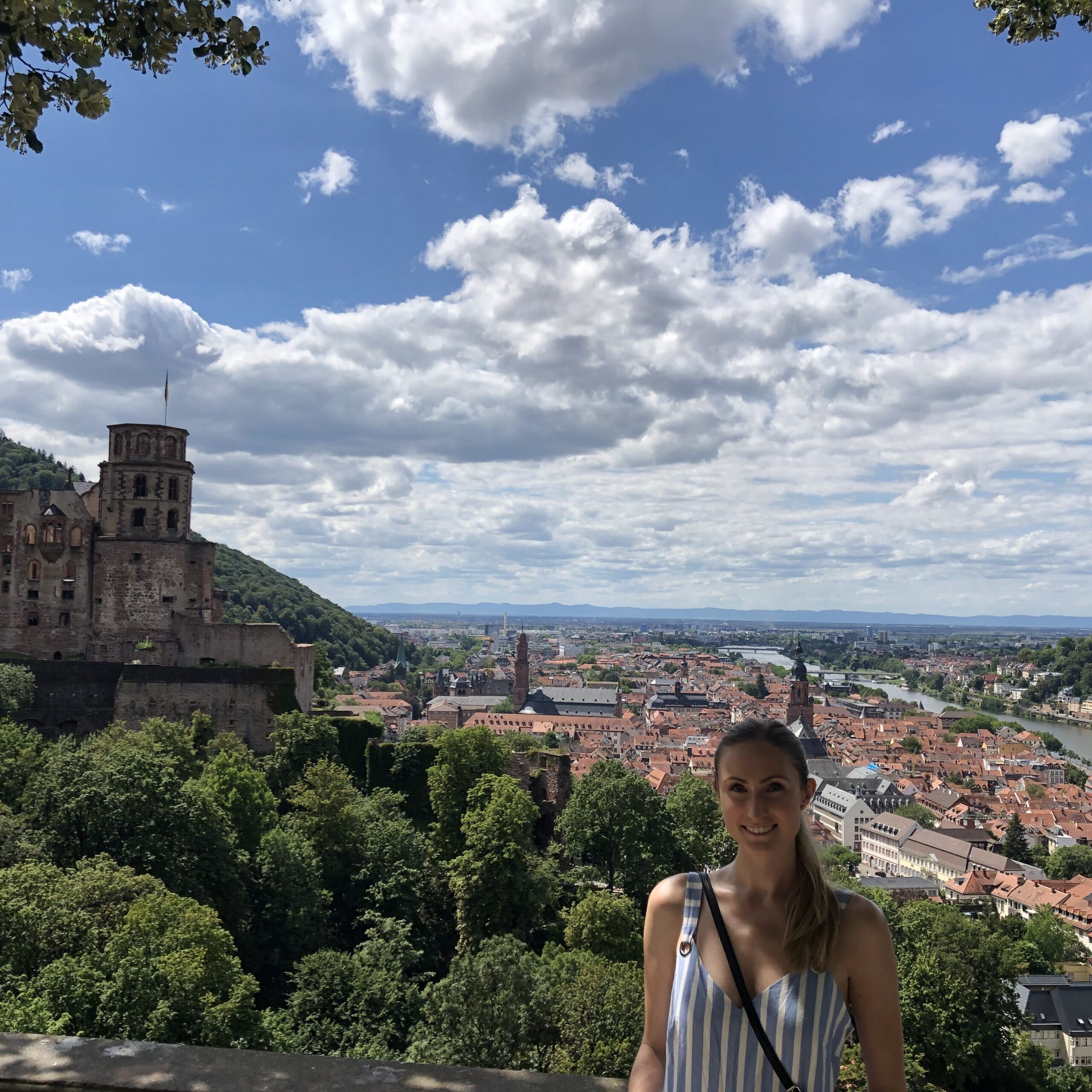 Person standing at an overlook above a historic hilltop castle and a riverfront city, with green hills and a sky filled with large white clouds in the background
