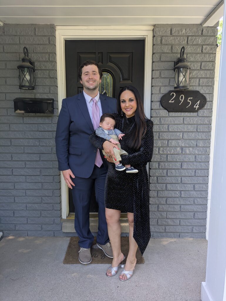Adult couple standing with an infant outside a brick home.