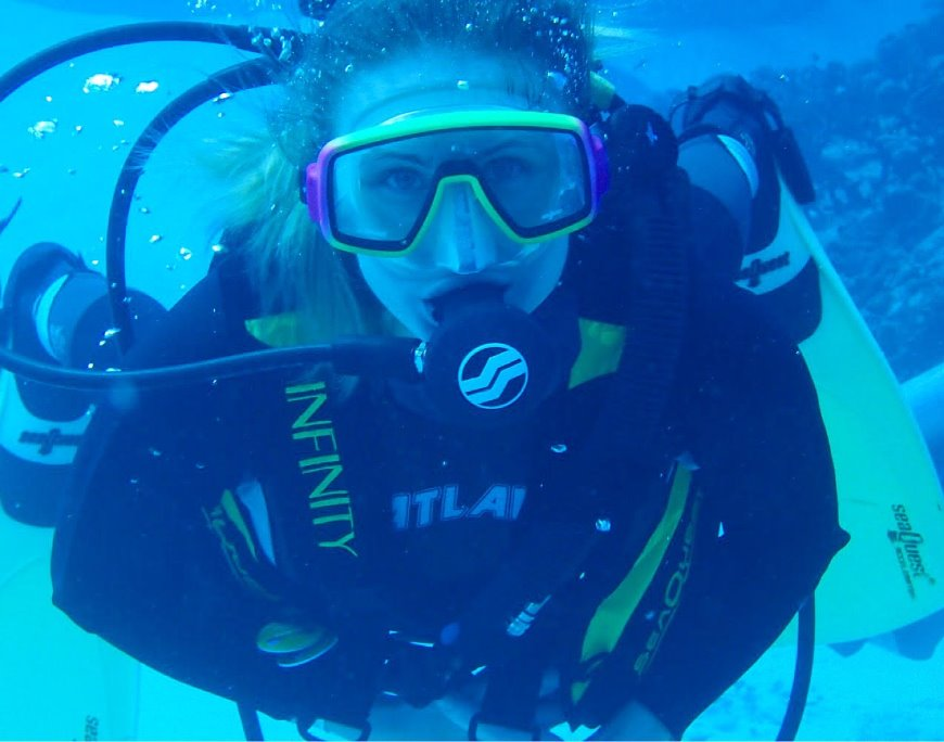 Scuba diver underwater wearing a mask, regulator, and air tank, swimming near a coral reef in clear blue water