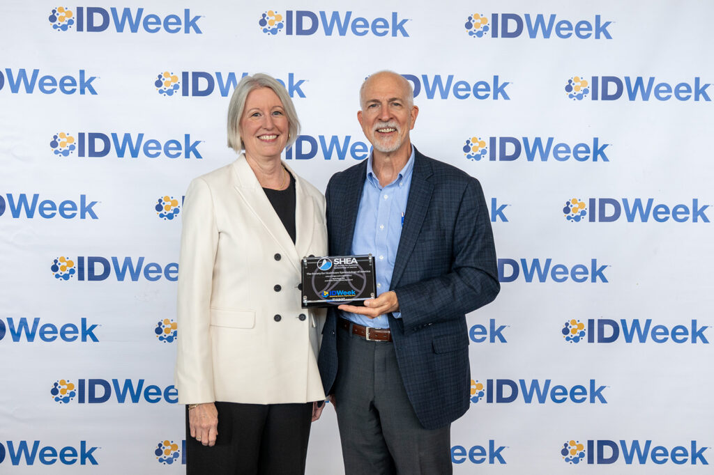 Two individuals pose in front of an IDWeek step-and-repeat backdrop while holding a plaque recognizing an achievement or award. The plaque displays SHEA and IDWeek logos.