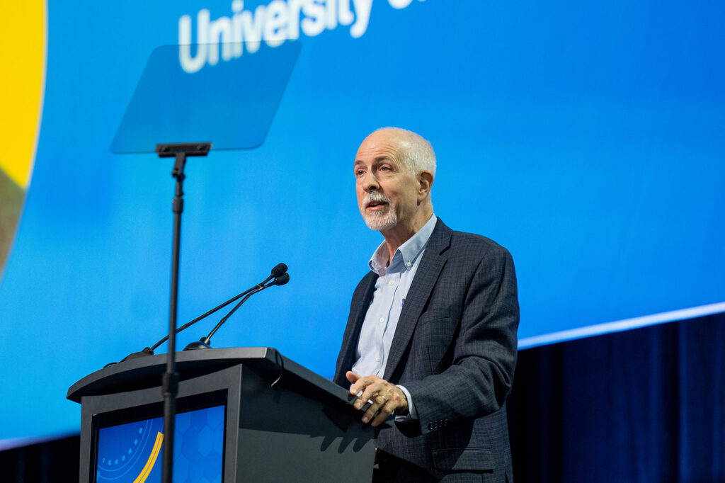 Speaker addressing an audience from a podium at an IDWeek conference session. A teleprompter stands in front of the podium, and a large blue presentation screen forms the background.