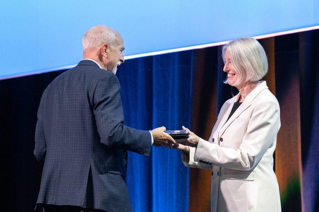 Award presentation on a conference stage, with two individuals exchanging a commemorative plaque before a blue-lit backdrop.
