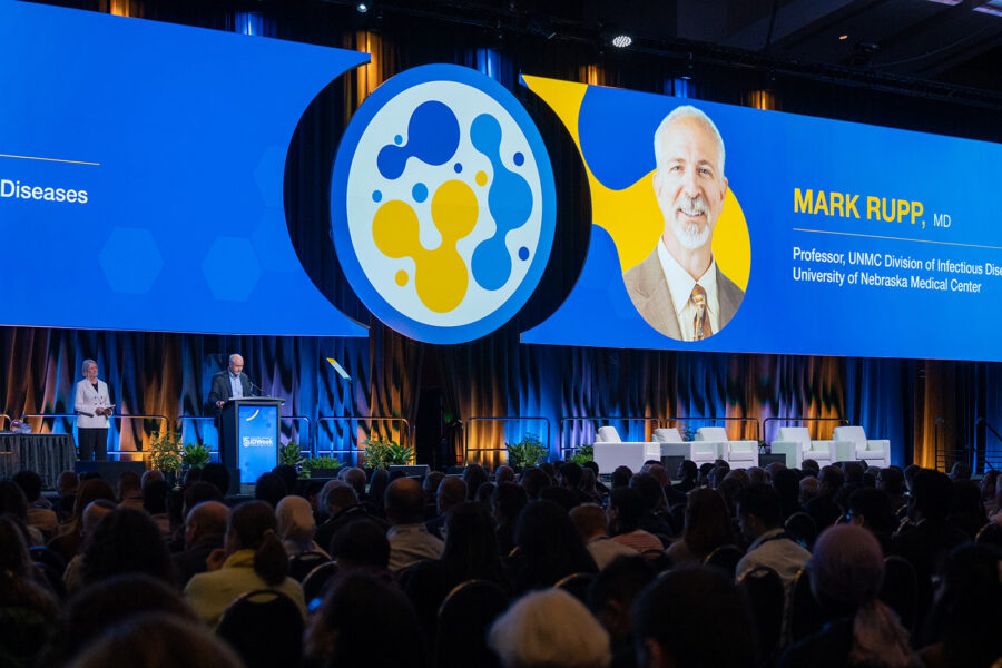 An IDWeek conference stage featuring a large presentation screen with a portrait and the text identifying Mark Rupp, MD, as Professor in the UNMC Division of Infectious Diseases at the University of Nebraska Medical Center. One individual speaks at the podium while another stands nearby on stage.