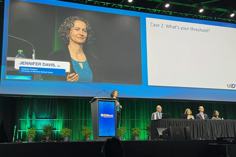 A speaker presents from the podium during an IDWeek session while a large projection screen displays the presentation title slide reading “Case 2: What’s your threshold?” Four panelists are seated at a table on stage, and the IDWeek logo appears on the podium.