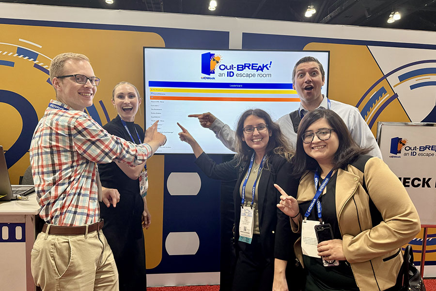 Five conference attendees wearing IDWeek badges stand in front of an interactive “Out-BREAK! An ID escape room” display at the IDWeek exhibit hall. The group points toward a leaderboard shown on a large digital screen, with IDWeek branding and a blue-and-gold exhibit backdrop visible behind them.