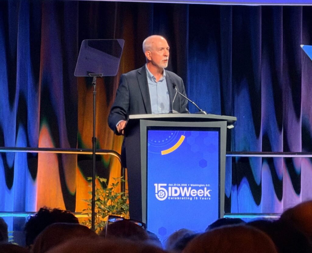 A speaker stands behind an IDWeek-branded podium on a large conference stage illuminated in blue and gold lighting. Teleprompter screens flank the podium, and audience members are visible in the foreground.