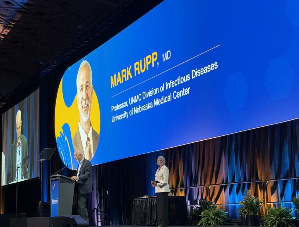 A wide-angle view of an IDWeek conference session showing a full audience facing a large stage. The screen displays a recognition slide for Mark Rupp, MD, Professor in the UNMC Division of Infectious Diseases at the University of Nebraska Medical Center, while two individuals stand on stage beneath the screen.