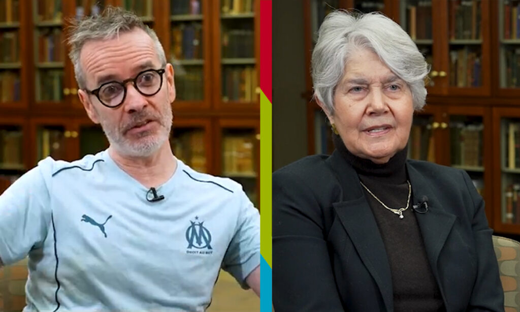 Man on left wearing black glasses and blue shirt. Woman with short silver hair on right wearing navy suit and black turtleneck. Bookshelves filled with books in the background