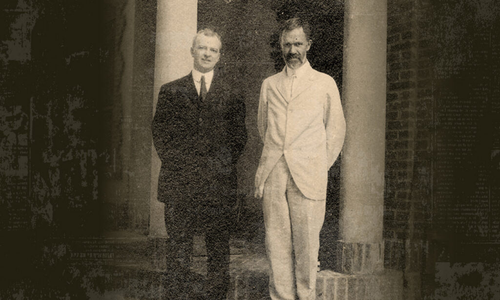 Faded image of two men in front of building columns. Man on left in black suite, man on right in white suite.