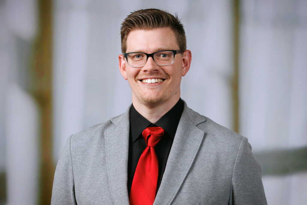 Portrait of man wearing glasses, black shirt, red tie and grey suit jacket.