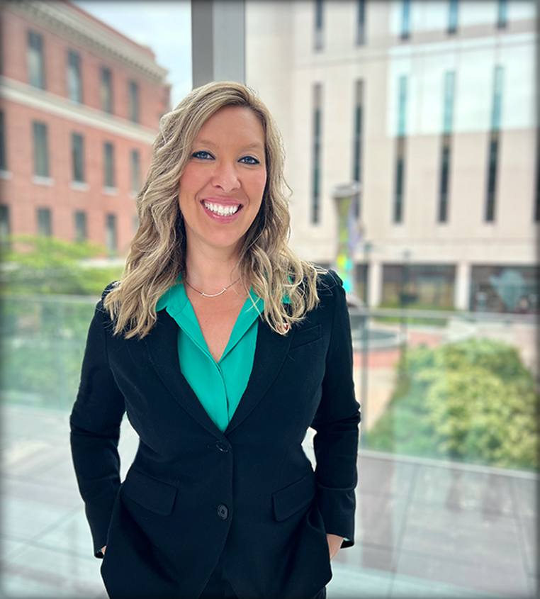 Woman in navy suit with teal shirt standing in front of large windows.